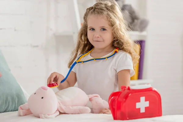 Adorable niño examinando conejo juguete con estetoscopio en habitación de los niños y mirando a la cámara - foto de stock