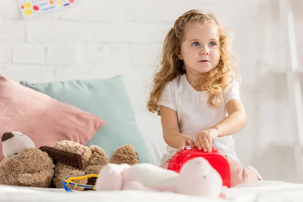 Adorable kid opening first aid kit, rabbit toy on bed in children room — Stock Photo