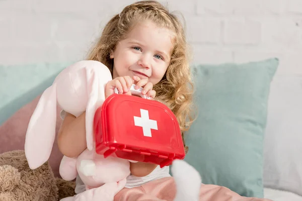 Criança adorável segurando kit de primeiros socorros e brinquedo de coelho na sala de crianças, olhando para a câmera — Fotografia de Stock