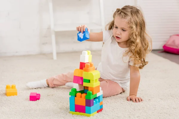 Adorable niño con pelo rizado jugando con el constructor de plástico de color en la alfombra en la habitación de los niños - foto de stock