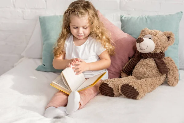 Adorable child playing with teddy bear and reading book on bed in children room — Stock Photo