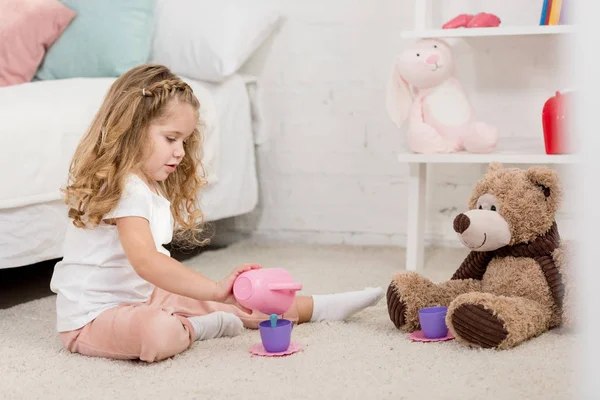 Adorabile bambino che gioca con orsacchiotto e bicchieri di plastica sul pavimento nella stanza dei bambini — Foto stock