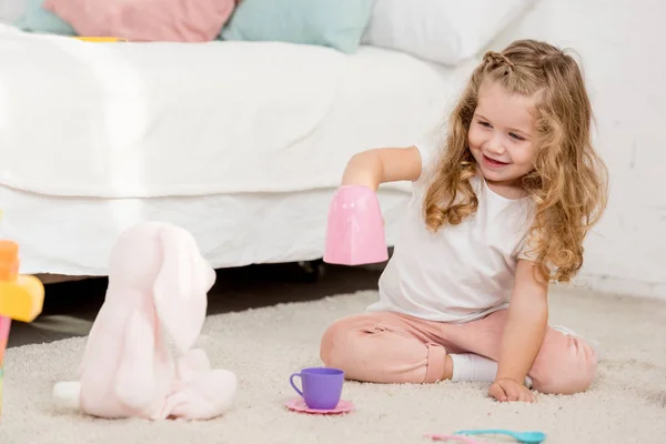 Adorable feliz niño jugando con conejo juguete y vasos de plástico en la habitación de los niños - foto de stock