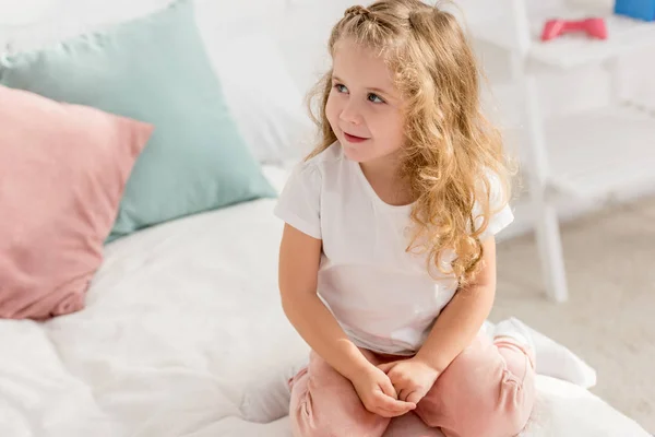 Vista de ángulo alto de adorable niño sentado en la cama en la habitación de los niños y mirando hacia otro lado - foto de stock