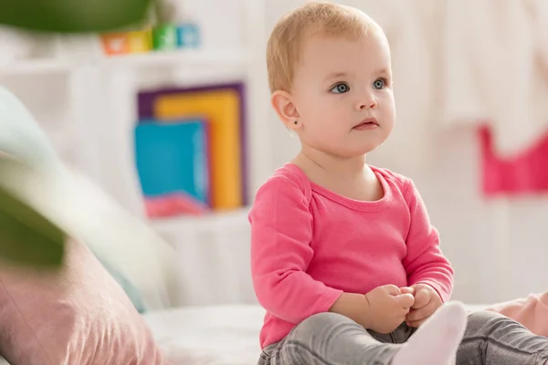 Прелестный малыш в розовой рубашке сидит на кровати и смотрит вдаль в детской комнате — стоковое фото