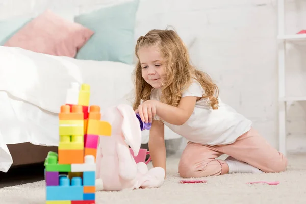 Criança adorável com cabelo encaracolado brincando com coelho macio e copos de plástico no tapete na sala de crianças — Fotografia de Stock