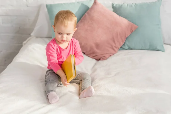 Adorable kid in pink shirt holding book on bed in children room — Stock Photo