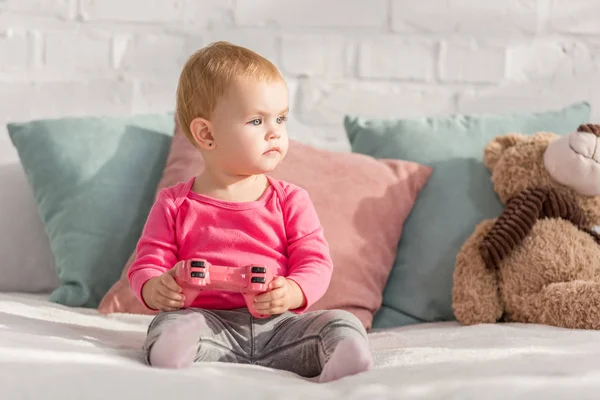 Adorable kid in pink shirt holding pink joystick on bed in children room and looking away — Stock Photo