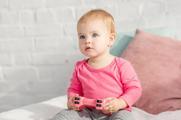 Adorable kid in pink shirt holding pink joystick on bed in children room and looking up — Stock Photo