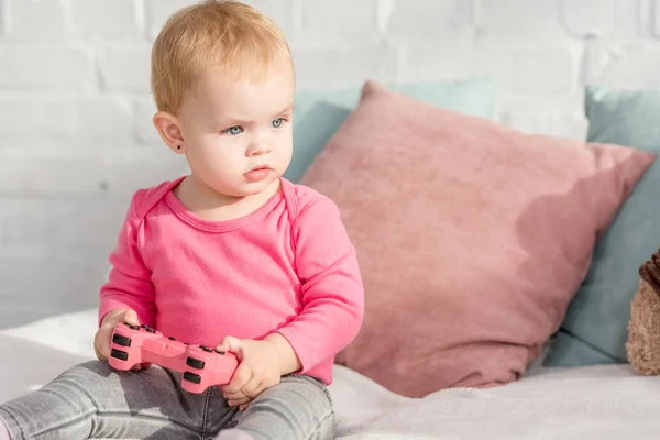 Adorable kid in pink shirt holding pink gamepad on bed in children room — Stock Photo