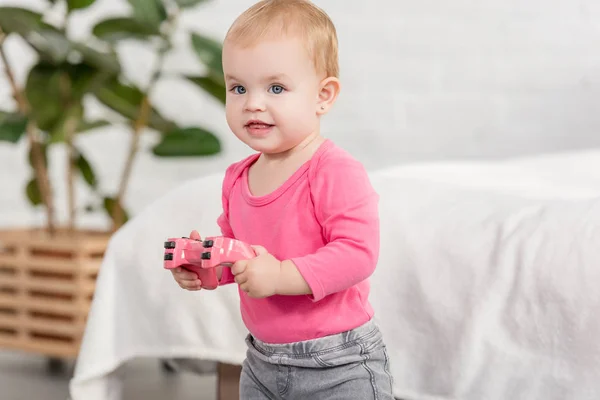 Criança adorável alegre na camisa rosa segurando joystick rosa perto da cama no quarto das crianças — Fotografia de Stock