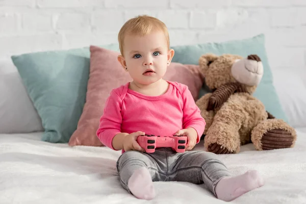 Adorable child in pink shirt holding pink joystick on bed in children room — Stock Photo