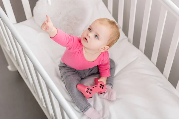 High angle view of adorable kid in pink shirt holding pink joystick in crib and pointing on toy — Stock Photo