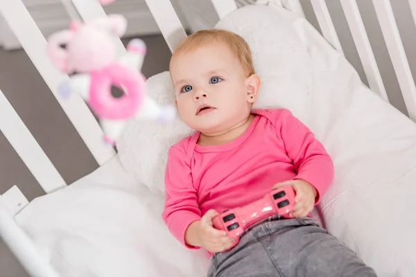 High angle view of adorable kid in pink shirt holding pink joystick and lying in crib — Stock Photo