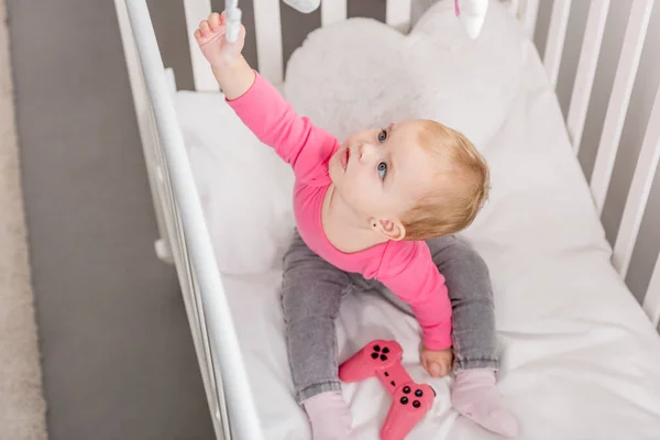 High angle view of adorable kid in pink shirt holding pink joystick in crib and reaching toy — Stock Photo