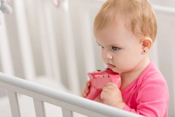 Adorable kid in pink shirt holding pink joystick in crib and looking away — Stock Photo