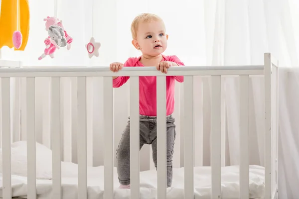 Adorable kid in pink shirt standing in crib and looking away — Stock Photo