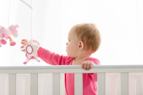 Side view of adorable kid in pink shirt standing in crib and touching toys — Stock Photo