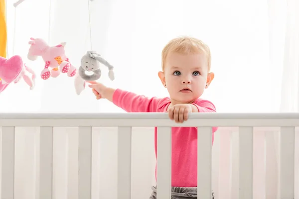 Adorable kid in pink shirt standing in crib and touching toys and looking up — Stock Photo