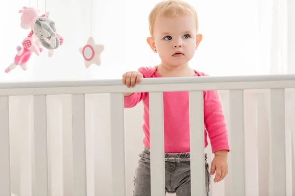 Adorable niño en camisa rosa de pie en cuna y mirando hacia otro lado - foto de stock