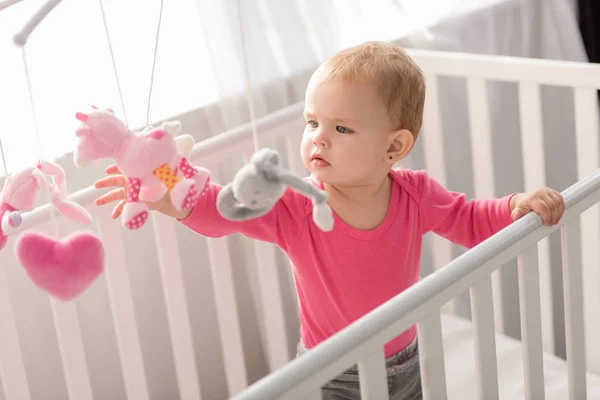 Adorable child in pink shirt standing in crib and touching toys — Stock Photo
