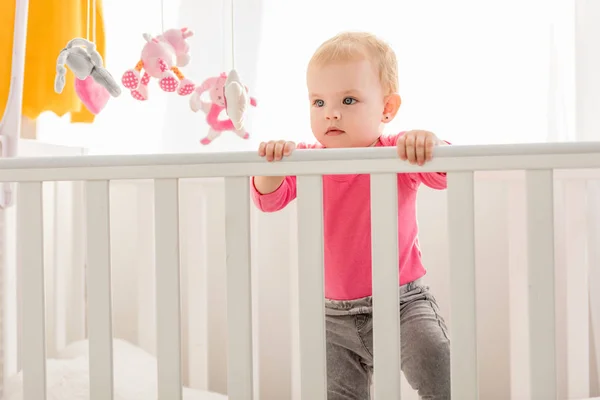 Очаровательный ребенок в розовой рубашке стоит в кроватке и смотрит в сторону — стоковое фото