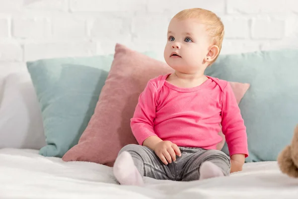 Adorable kid sitting on bed with pillows and looking up in children room — Stock Photo