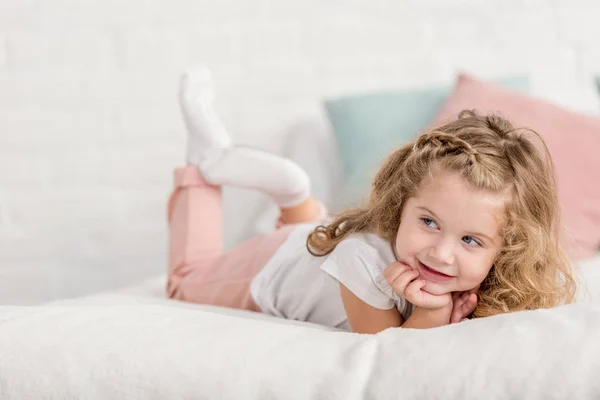 Fröhlich liebenswertes glückliches Kind, das im Kinderzimmer auf dem Bett liegt und wegschaut — Stockfoto