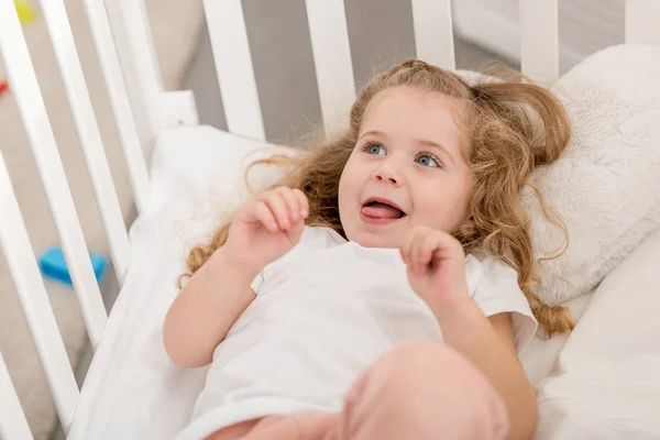 Adorable kid sticking tongue out and lying in crib in children room — Stock Photo