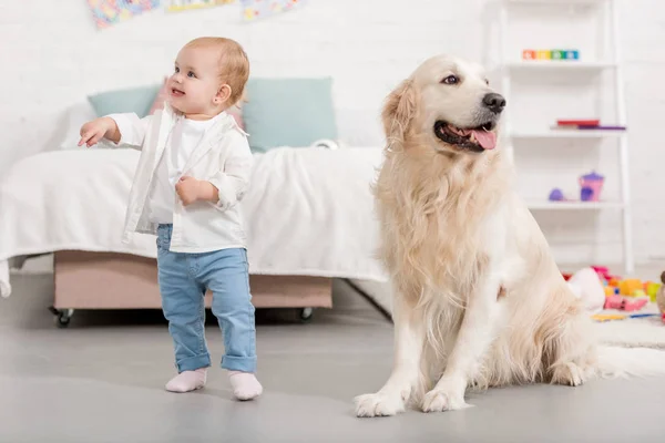 Niño adorable feliz mirando hacia otro lado, golden retriever sentado en el suelo en la habitación de los niños - foto de stock