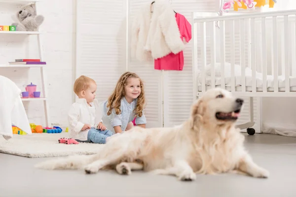Selective focus of cheerful adorable sisters playing on floor, golden retriever lying near in children room — Stock Photo