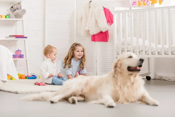 Enfoque selectivo de hermanas adorables alegres jugando en el suelo, golden retriever acostado cerca en la habitación de los niños - foto de stock