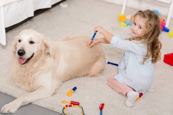 Vue grand angle d'adorable enfant faisant semblant vétérinaire et examinant golden retriever dans la chambre des enfants — Photo de stock