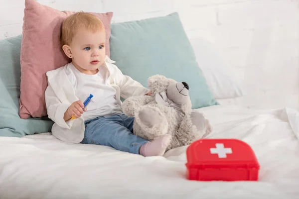 Adorable niño sentado en la cama con osito de peluche y botiquín de primeros auxilios en habitación para niños - foto de stock