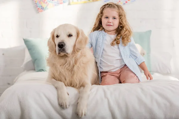 Adorable niño y golden retriever perro sentado en la cama en la habitación de los niños - foto de stock