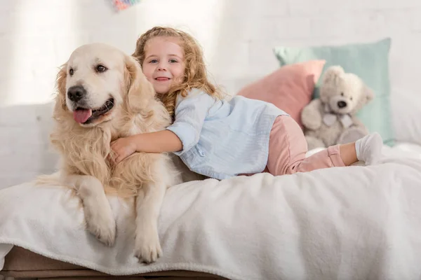 Adorável criança feliz e golden retriever deitado na cama no quarto das crianças — Fotografia de Stock