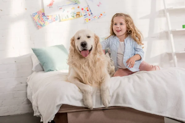 Adorable niño y mullido golden retriever sentado en la cama juntos en la habitación de los niños - foto de stock