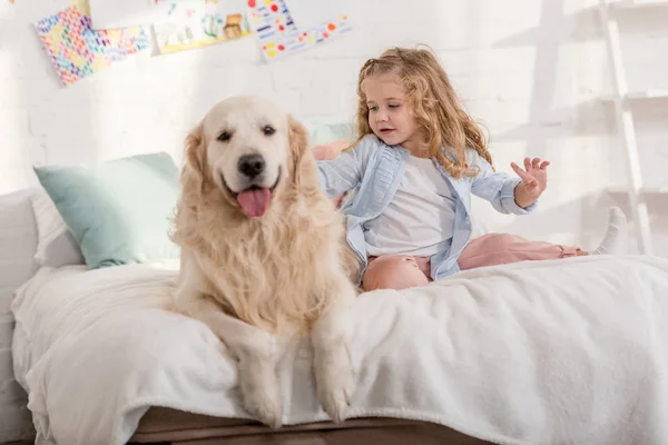 Очаровательный ребенок ласкает золотистого ретривера на кровати вместе в детской комнате — стоковое фото