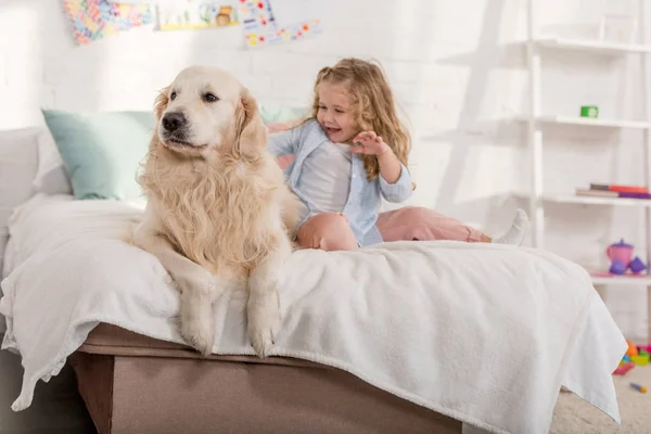 Happy adorable kid palming golden retriever on bed together in children room — Stock Photo