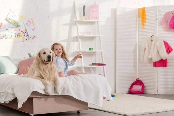 Entzückendes Kind und Golden Retriever sitzen zusammen auf dem Bett im Kinderzimmer, Kind zeigt auf etwas — Stockfoto