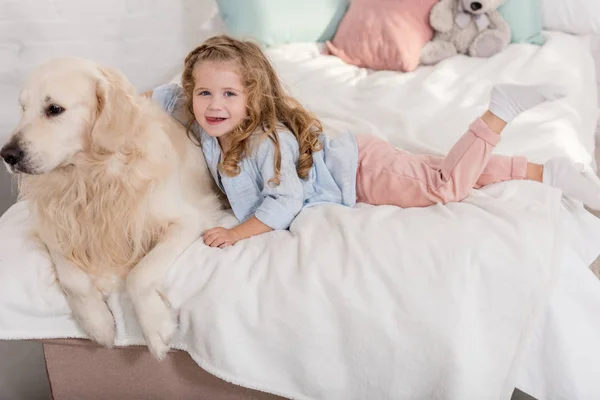 Vista de ángulo alto de adorable niño abrazando golden retriever en la cama en la habitación de los niños y mirando a la cámara - foto de stock