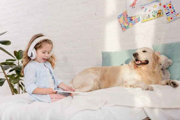 Чарівна дитина слухає музику з планшетом, золотий ретривер лежить на ліжку в дитячій кімнаті — стокове фото
