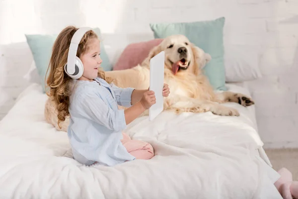 Happy adorable kid listening music with tablet, golden retriever lying on bed in children room — Stock Photo