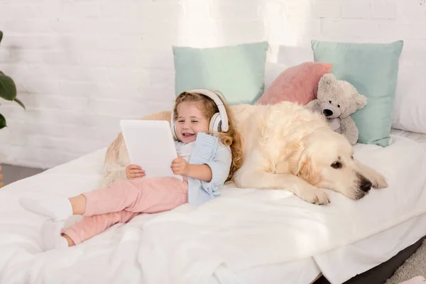 Sorridente adorabile bambino che ascolta musica con tablet e si appoggia sul golden retriever sul letto nella stanza dei bambini — Foto stock