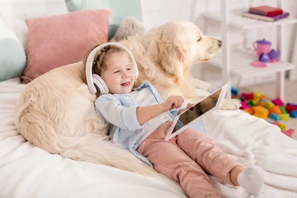 Adorável criança alegre ouvir música com tablet e inclinando-se em golden retriever na cama na sala de crianças — Fotografia de Stock