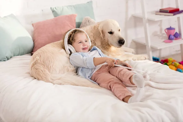 Adorable kid listening music with tablet and leaning on fluffy golden retriever on bed in children room — Stock Photo