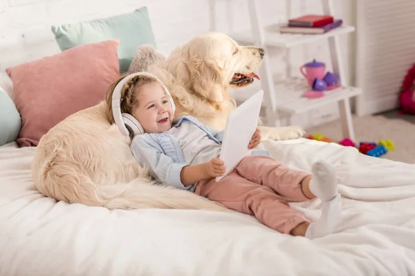 Sorridente adorabile bambino che ascolta musica con tablet e si appoggia sul golden retriever sul letto nella stanza dei bambini — Foto stock