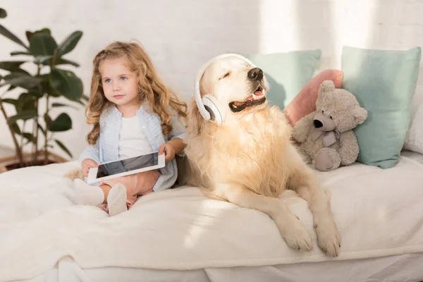 Adorable kid holding tablet, golden retriever with headphones lying on bed in children room — Stock Photo