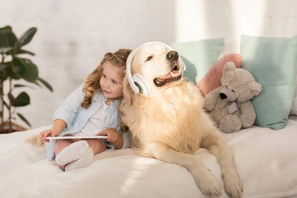 Adorável criança segurando tablet, bonito golden retriever com fones de ouvido deitado na cama na sala de crianças — Fotografia de Stock