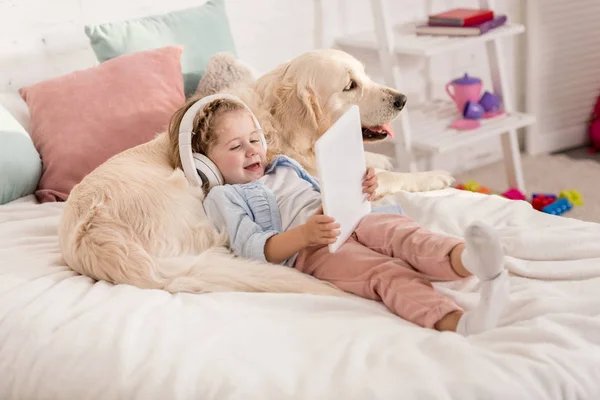 Adorable niño feliz escuchando música con la tableta y apoyándose en perro retriever dorado en la cama en la habitación de los niños - foto de stock